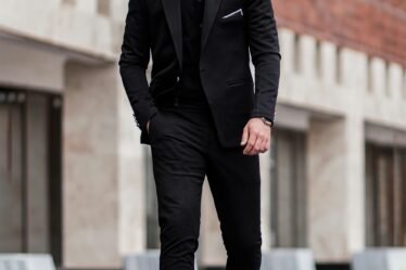 homme en veste de costume noir et pantalon noir debout sur un sol en beton gris pendant la journee